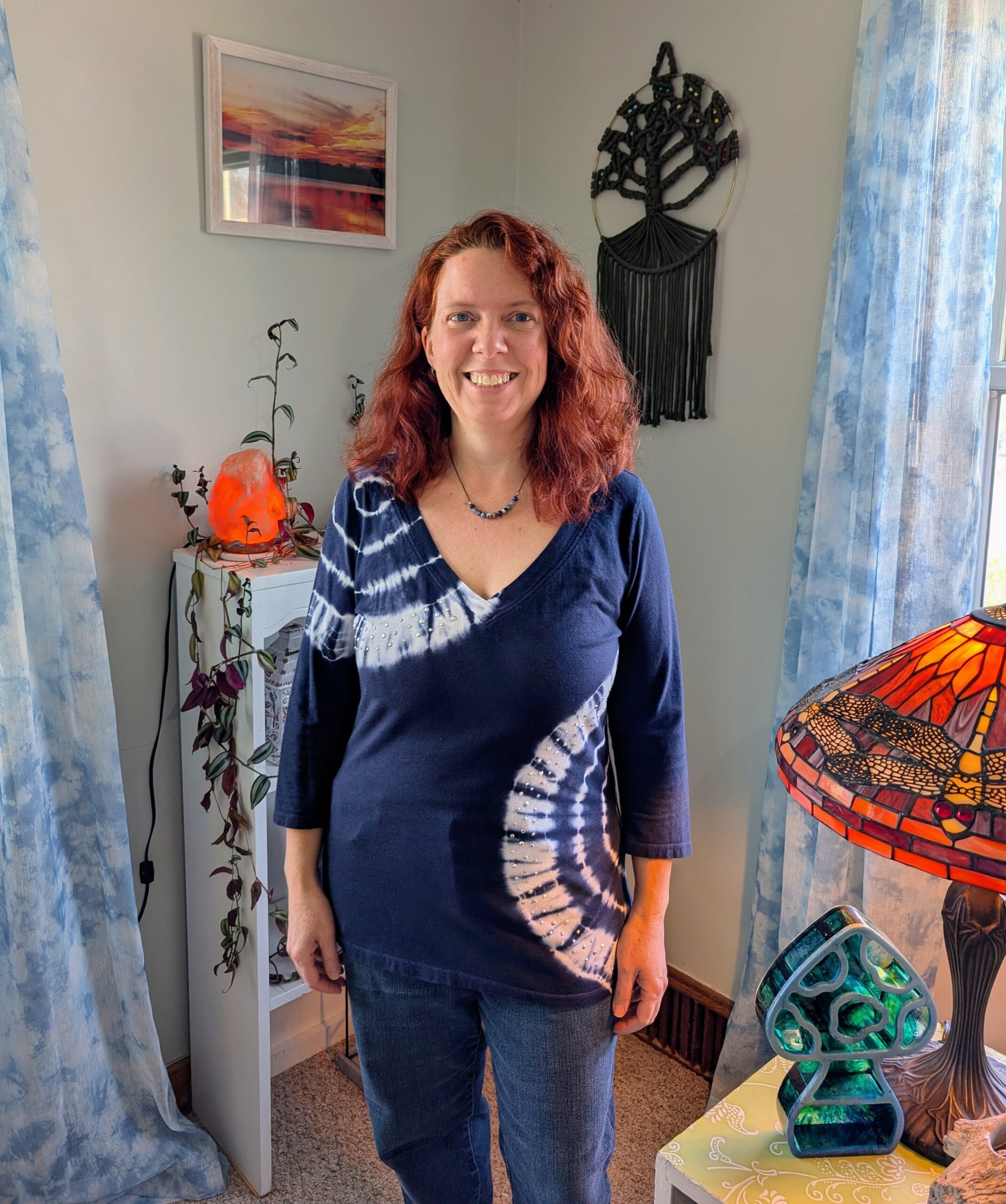Amanda Dacio with wavy red hair, wearing a blue tie-dye shirt and jeans, stands smiling in a bright room with blue curtains, plants, a decorative stained glass lamp, and wall art featuring a tree and a sunset.
