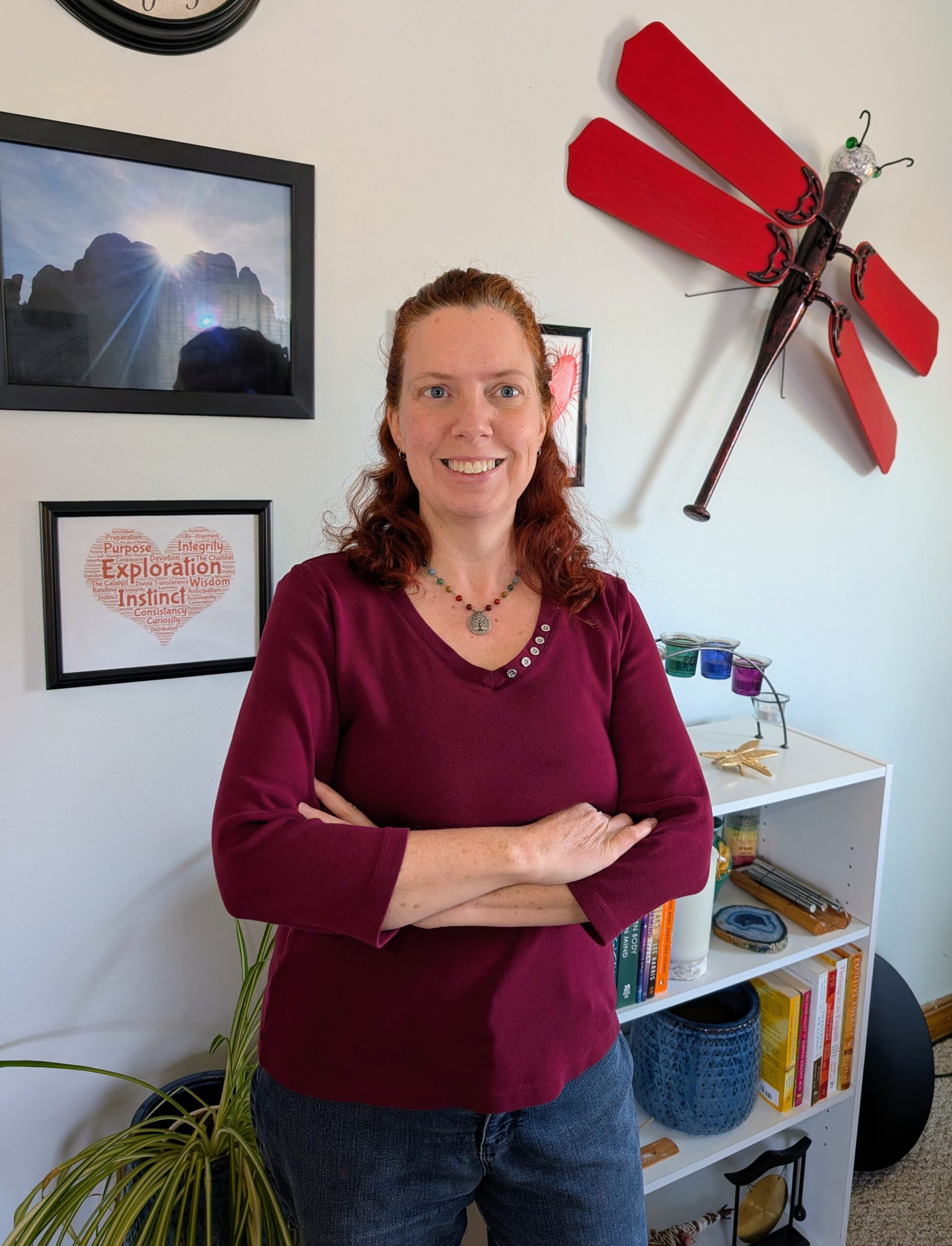 Amanda Dacio with long red hair, wearing a maroon top and jeans, stands smiling with arms crossed in a cozy room. Behind her are shelves with books and decor, framed photos, and a large red dragonfly wall decoration.